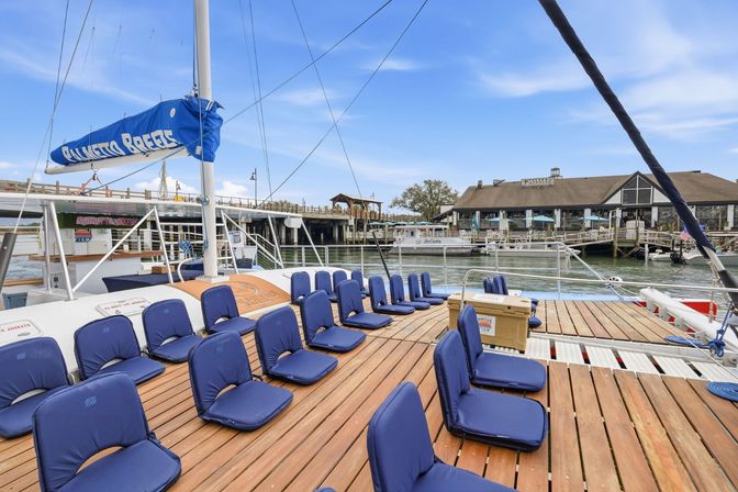 Wooden sailboat deck lined with blue cushioned seats, docked at a sunny marina with waterfront restaurants, piers, and boats in the background.