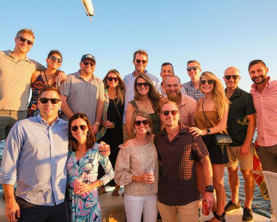 Group of friends on a sunset boat cruise, smiling in sunglasses and summer outfits, holding drinks with golden-hour ocean backdrop.