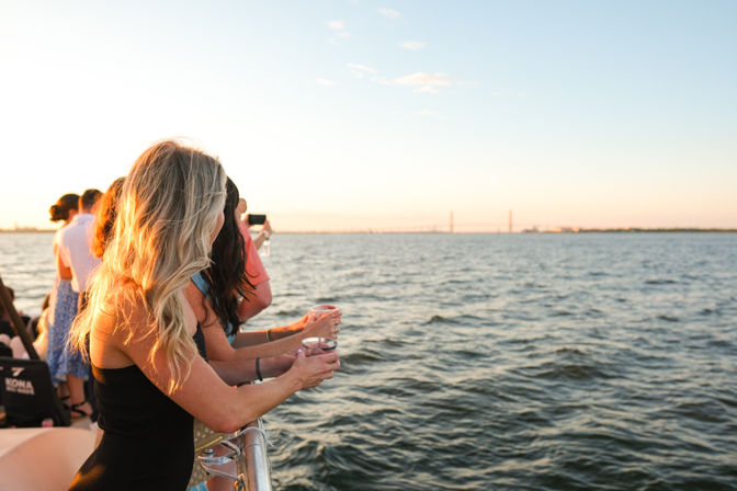 Group of people on a sunset boat cruise leaning on the railing with drinks, watching a distant bridge across calm coastal waters in golden-hour light.