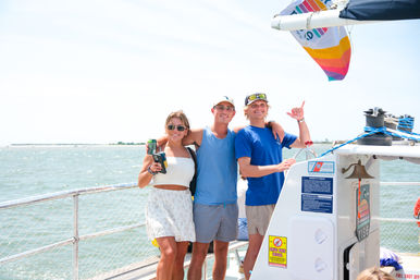 Three friends smiling on a sunlit sailboat deck near the coastline, one at the helm and two holding drinks during a cheerful summer sailing trip on calm coastal waters.