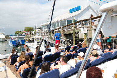 Boat tour passengers seated on a sunny marina deck listening to a guide gesturing on the dock beside a waterfront restaurant with moored yachts in the harbor.