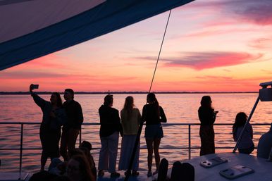 Silhouetted group on a boat railing watching a vibrant pink-orange sunset over calm water during a peaceful sunset cruise