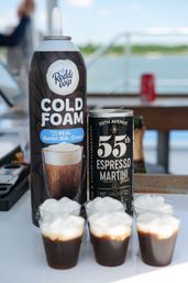 Can of cold foam topping next to a canned espresso martini on a boat deck by the water, with six small espresso-martini shots in plastic cups topped with white foam.