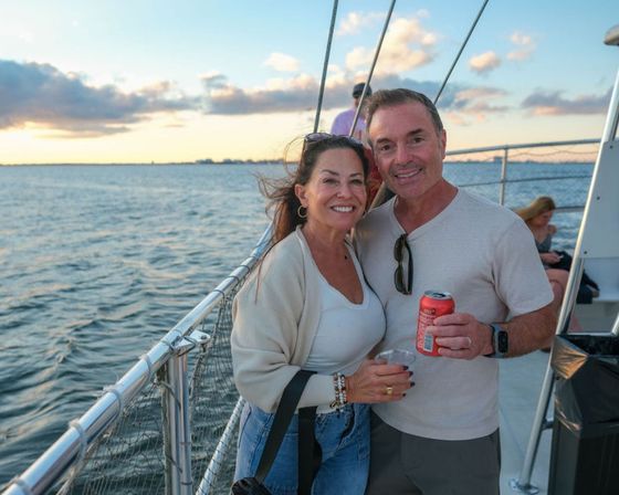 Smiling couple on a sunset cruise, standing at the boat railing with drinks in hand, calm ocean and distant coastline under a colorful sky.