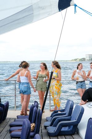 Group of young women chatting and laughing on a sailboat deck with blue cushioned seats, white sail overhead and coastal water in the background