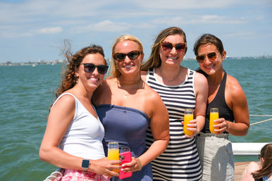 Four smiling women in sunglasses on a sunny summer boat cruise, holding mimosas by the railing with blue bay water and a distant shoreline under a partly cloudy sky.