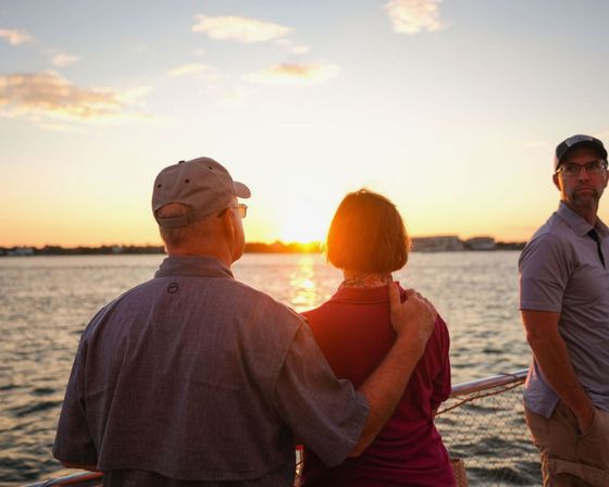 Sunset boat scene over calm water: two people standing arm‑in‑arm on the deck watching the sun as a third person glances back.