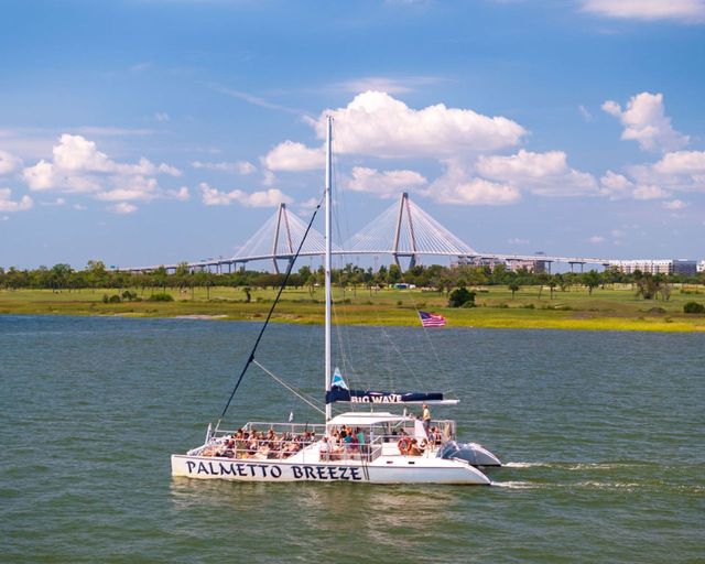 Catamaran sailboat with passengers cruising a calm coastal bay, American flag on the mast, marshy shoreline and a cable‑stayed bridge under a bright blue sky with puffy clouds.
