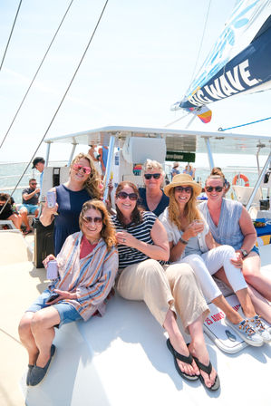 Cheery group of friends wearing sunglasses and a straw hat, enjoying drinks on a sunny catamaran sail with the ocean in the background.