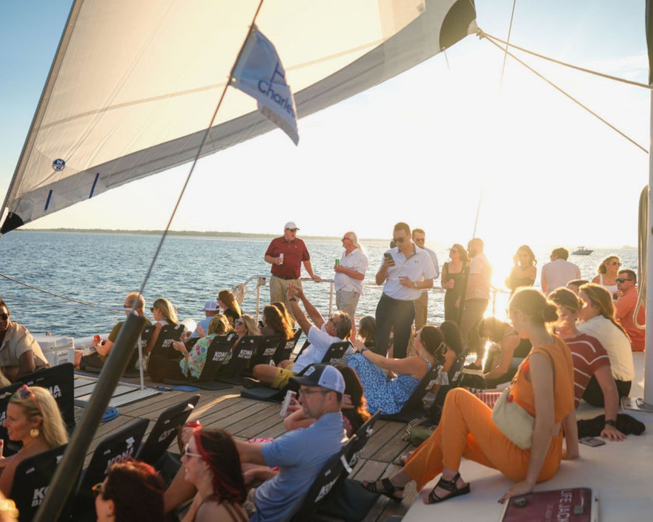 Sunset sail on calm coastal waters — a crowd of people lounging and socializing on a sailboat deck with raised sails, folding chairs, drinks, and a distant shoreline.