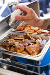 Catered brunch buffet: foil chafing pan filled with golden-brown French toast pieces and sweet pastries dusted with powdered sugar, a hand pouring syrup from a metal dispenser