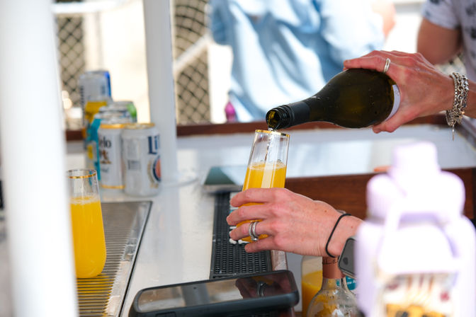 Hands pouring sparkling wine into a flute of orange juice to make a mimosa at an outdoor brunch bar, with beer cans and bottles on the counter.
