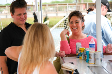 Three women chatting and smiling at a dockside boat bar, canned beers and a water bottle on the counter with marina railings in the background.