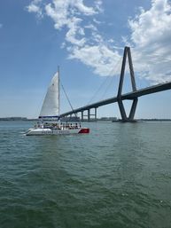 Tour catamaran with passengers sailing under a dramatic cable-stayed bridge in Charleston harbor on a sunny, partly cloudy day