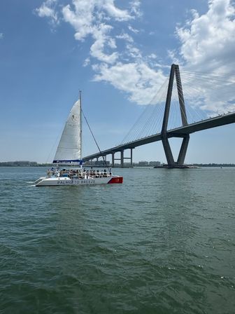 Tour catamaran with passengers sailing under a dramatic cable-stayed bridge in Charleston harbor on a sunny, partly cloudy day