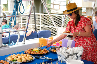 Woman in a straw hat and red dress serving appetizers from a buffet on a boat deck at a marina — platters of sliders, skewers and wrapped napkins on a blue tablecloth.