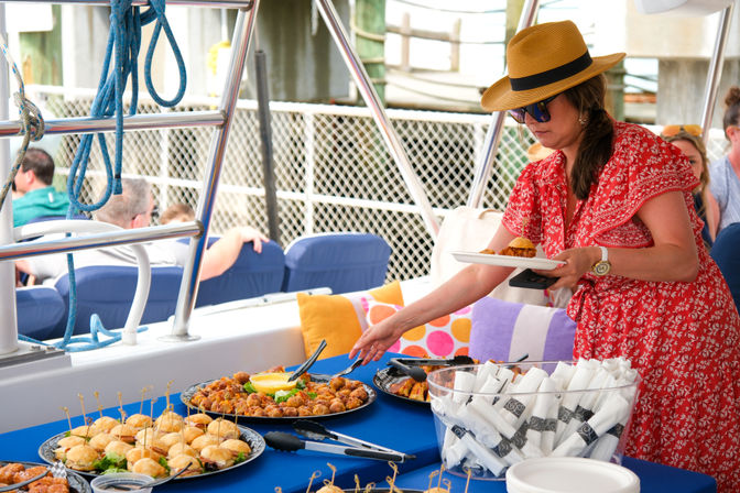 Woman in a straw hat and red dress serving appetizers from a buffet on a boat deck at a marina — platters of sliders, skewers and wrapped napkins on a blue tablecloth.