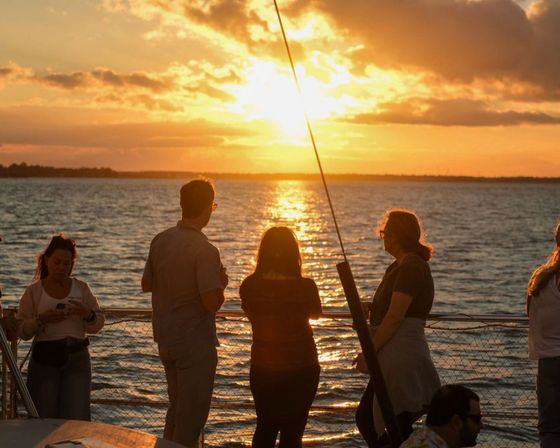 Group of people silhouetted on a sailboat deck watching a golden sunset reflect across calm coastal waters.