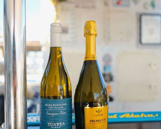 Two wine bottles on a bright bar counter: a Marlborough New Zealand Sauvignon Blanc with a screw cap and a foil-topped Prosecco sparkling wine in a casual indoor setting.