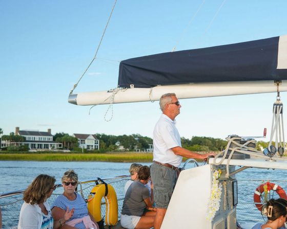 Leisure sailboat with a man at the helm and passengers relaxing as the boat cruises past marshy shoreline and waterfront homes on a sunny coastal inlet.