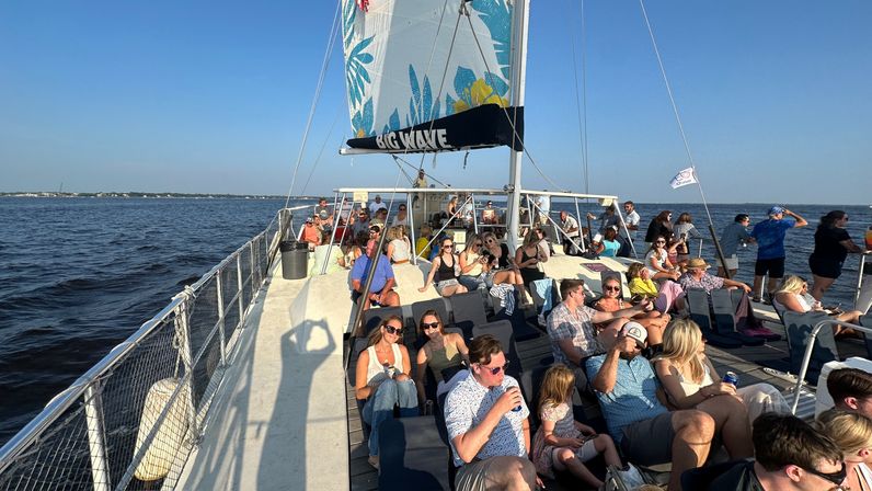 Sunlit group lounging on a catamaran cruise deck with a colorful sail overhead, passengers in summer clothes sipping drinks and enjoying calm coastal waters on a sunny afternoon.