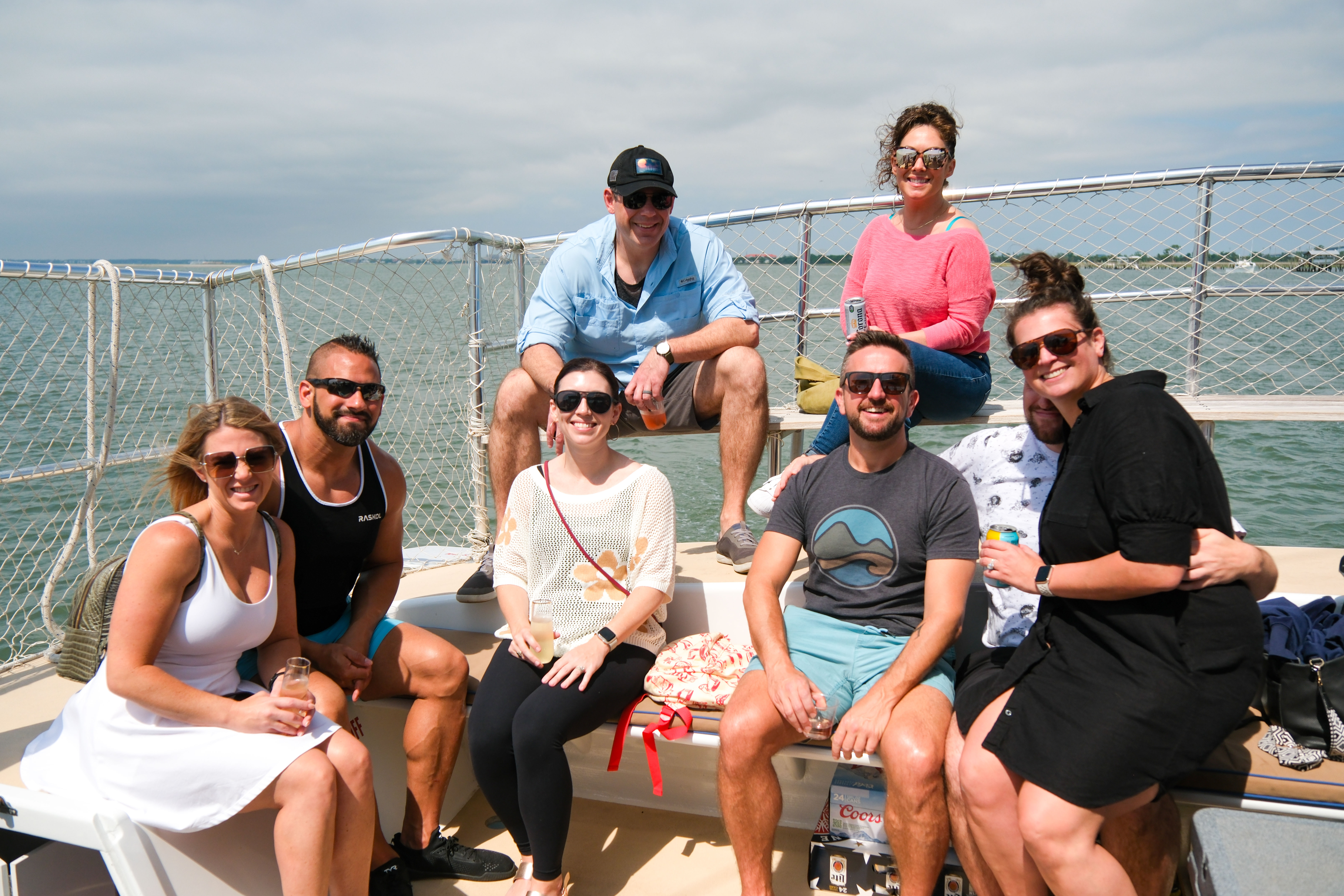 Eight friends smiling and posing on the deck of a leisure boat cruise in calm coastal waters, seated on benches with sunglasses and drinks under a bright, partly cloudy sky.
