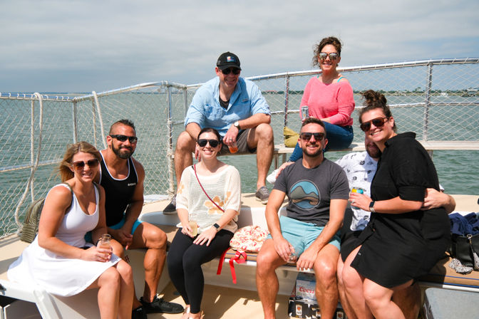 Eight friends smiling and posing on the deck of a leisure boat cruise in calm coastal waters, seated on benches with sunglasses and drinks under a bright, partly cloudy sky.