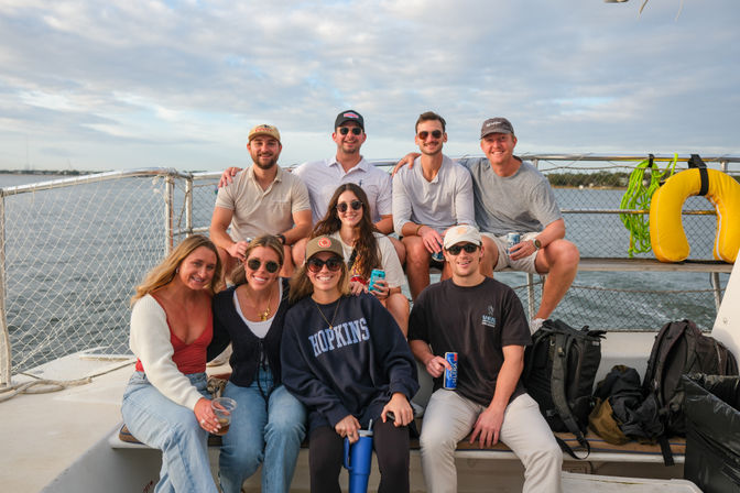 Smiling group of friends gathered on a boat deck during a coastal cruise, casually dressed with drinks, backpacks and a yellow life ring against a cloudy sky and calm water.