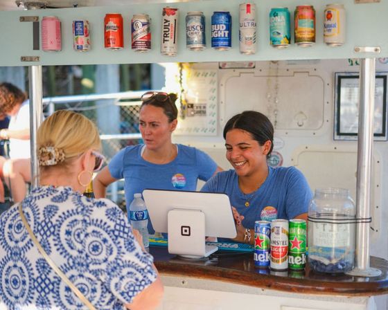 Two staff in blue shirts serving a customer at an outdoor beverage stand with a tablet POS on the counter, assorted beer and seltzer cans displayed overhead, and a tip jar visible.