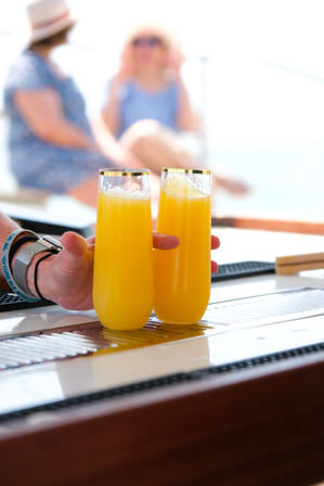 Two bright orange mimosas in tall flutes with gold rims on a yacht bar counter, a hand reaching for them and sunlit, blurred passengers on the deck in the background