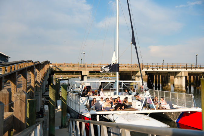 Group of people relaxing on a charter catamaran at a sunny marina dock beside a wooden boardwalk and low concrete bridge under a blue sky