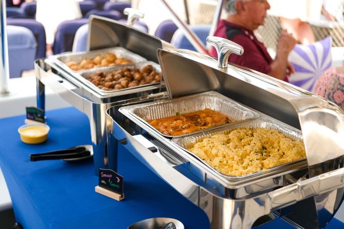 Catering buffet with shiny stainless steel chafing dishes on a blue tablecloth holding couscous, shrimp in sauce and seasoned meatballs, ready to serve at an event.