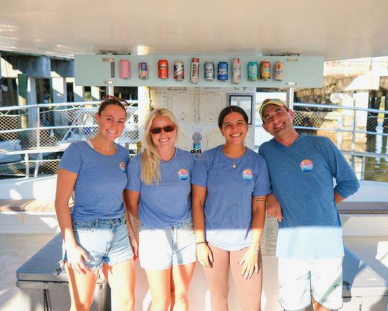 Smiling boat crew of four in matching blue shirts posing on a sunny coastal marina boat with canned drinks lined overhead and dock pilings in the background.