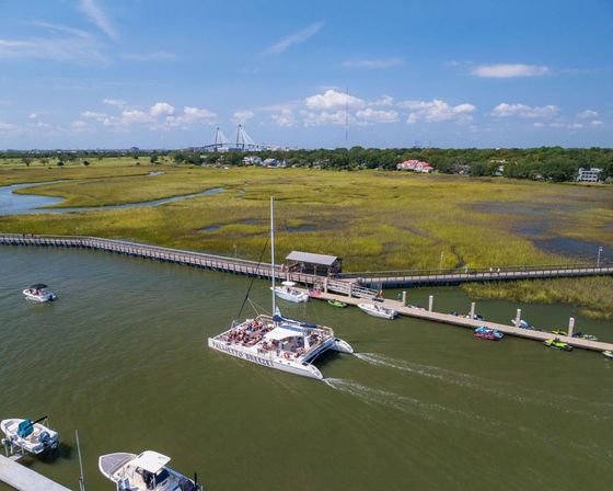 Aerial view of a busy catamaran tour and small boats moored by a wooden dock along golden tidal marshes, with a cable-stayed bridge and coastal homes under a bright blue sky.