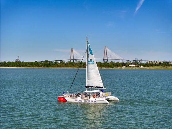 White catamaran sailboat with passengers gliding across calm coastal water on a sunny day, cable-stayed bridge and tree-lined shoreline in the background.