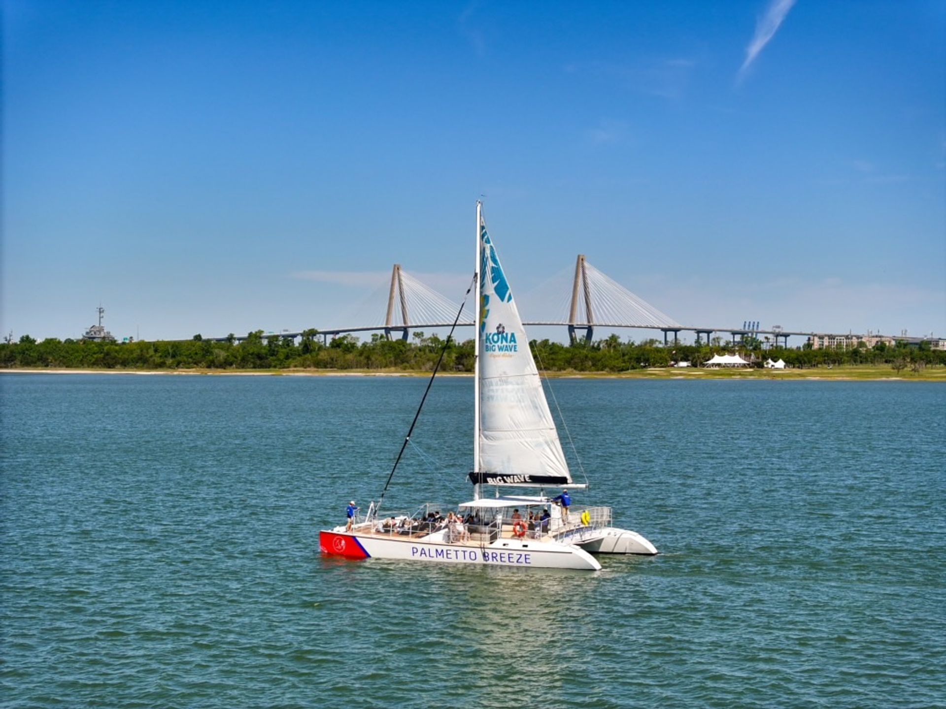 White catamaran sailboat with passengers gliding across calm coastal water on a sunny day, cable-stayed bridge and tree-lined shoreline in the background.