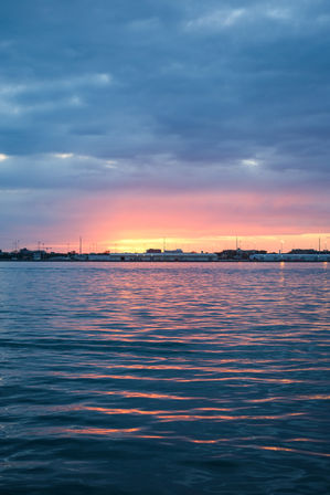 Pink and orange sunset over a calm harbor with blue clouded sky, rippling water reflecting warm hues and a distant shoreline of low buildings and lights