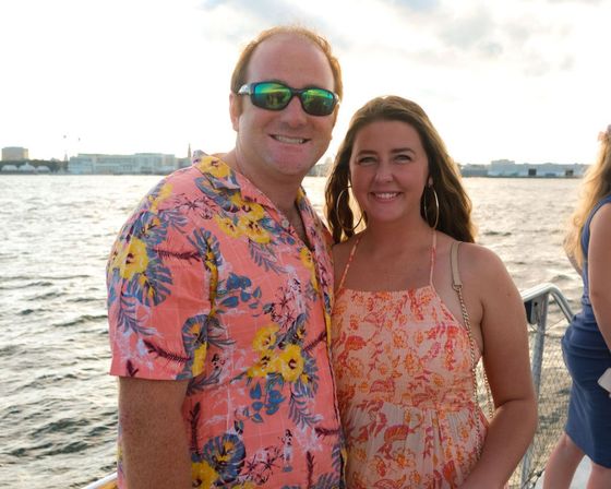 Smiling couple in a bright tropical shirt and floral sundress on a boat deck during a sunset harbor cruise with a city skyline across the water