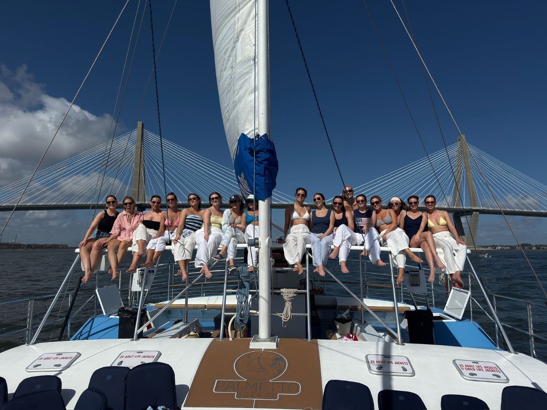 Group of people perched along the bow of a sailboat with legs dangling, sailing beneath a cable-stayed bridge on a sunny blue-sky day.