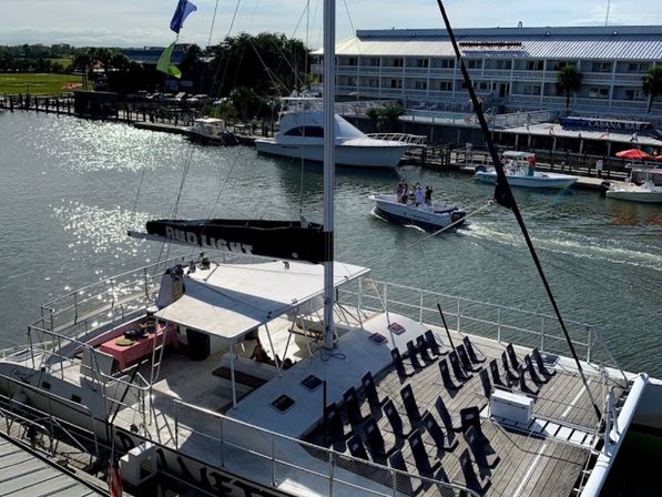 Sunlit coastal marina with a white catamaran decked in rows of navy lounge chairs, nearby yachts, a small powerboat cruising past waterfront docks.