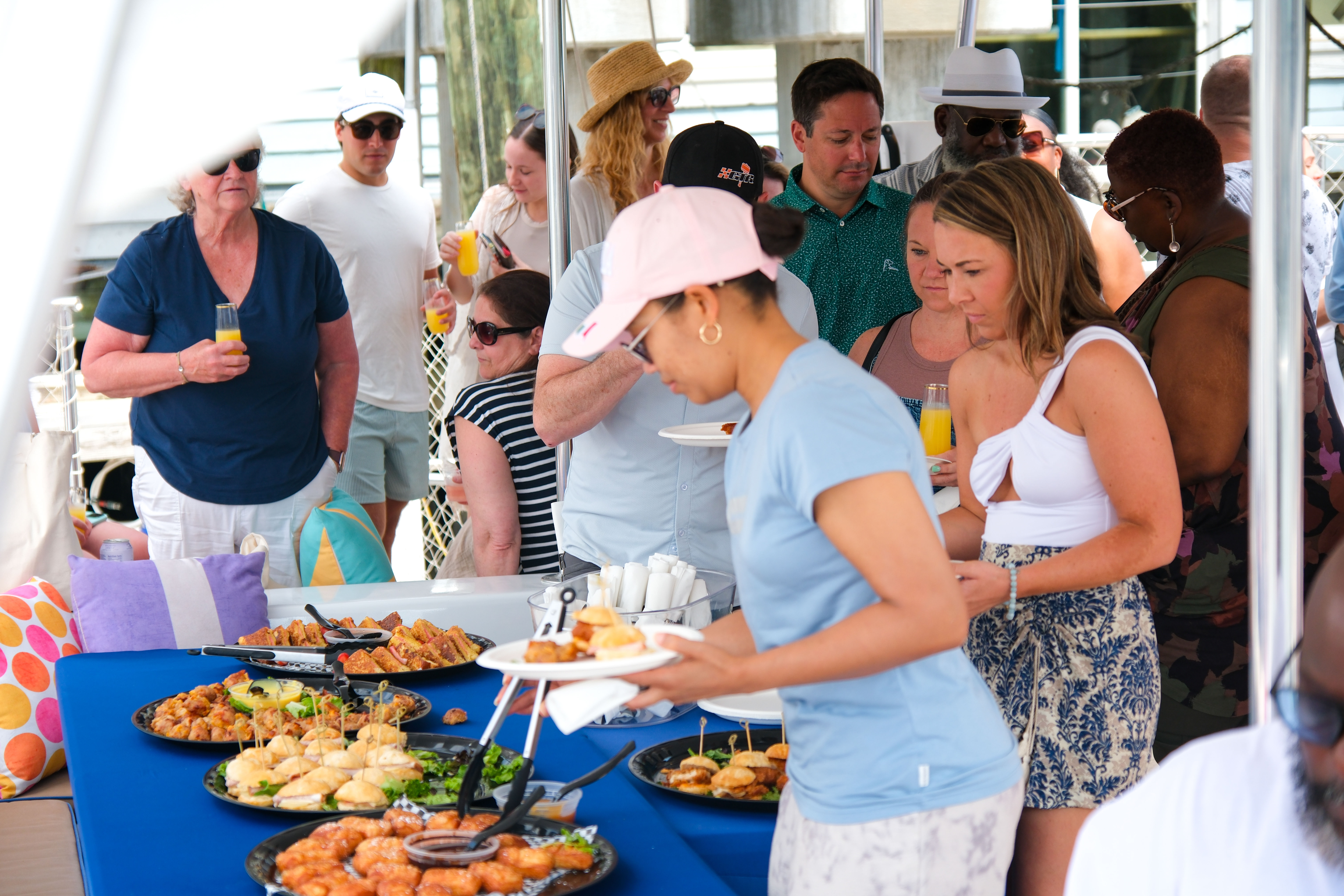 Guests grabbing sliders, pastries and appetizers from a blue-clothed buffet while holding mimosas at a sunny dockside brunch