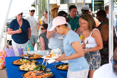 Guests grabbing sliders, pastries and appetizers from a blue-clothed buffet while holding mimosas at a sunny dockside brunch