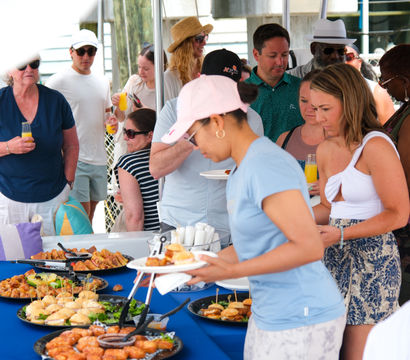 Guests grabbing sliders, pastries and appetizers from a blue-clothed buffet while holding mimosas at a sunny dockside brunch