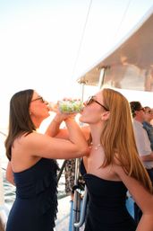 Two friends in navy dresses and sunglasses clink and drink canned beverages arm-in-arm aboard a sunlit boat during a sunset cruise on the ocean.