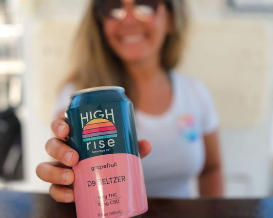 Close-up of hand holding a grapefruit D9 seltzer can labeled 5mg THC/10mg CBD, smiling woman in sunglasses blurred in a sunny outdoor background