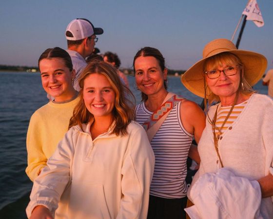 Group of four smiling women on a summer sunset boat ride, wearing casual layers and a sun hat with calm coastal water in the background