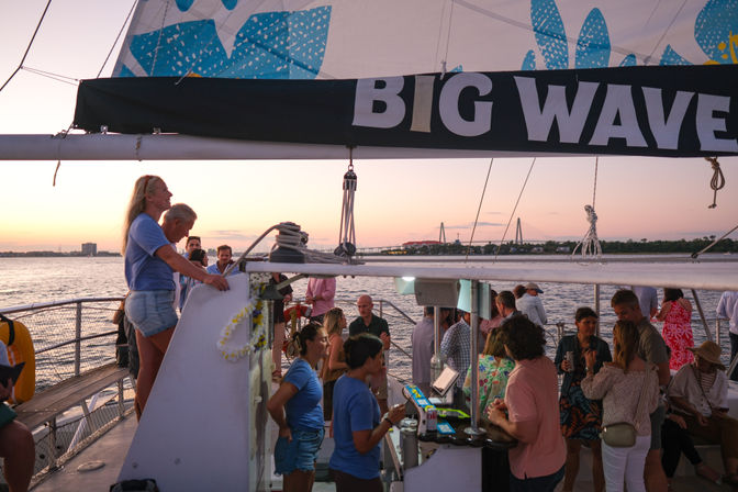 Sunset sailboat party in a harbor — crowded deck under a 'BIG WAVE' sail, people mingling with city skyline and suspension bridge on the horizon.
