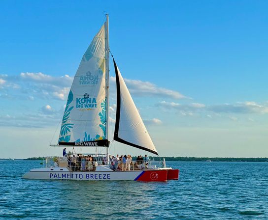 Catamaran sailboat with a colorful mainsail and a group of passengers cruising on calm blue coastal waters at golden hour.
