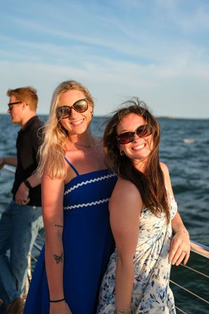 Two smiling friends in sunglasses on a boat at sea at golden hour — one in a blue sundress and one in a floral dress with ocean and sky in the background.