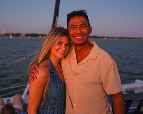 Smiling couple on a sailboat at sunset enjoying a coastal sunset sail with calm harbor waters and a distant shoreline in the background.